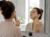 Comment faire un drainage lymphatique du visage soi-même ? Une femme pratique un drainage lymphatique visage avec un Gua Sha en jade devant un miroir.