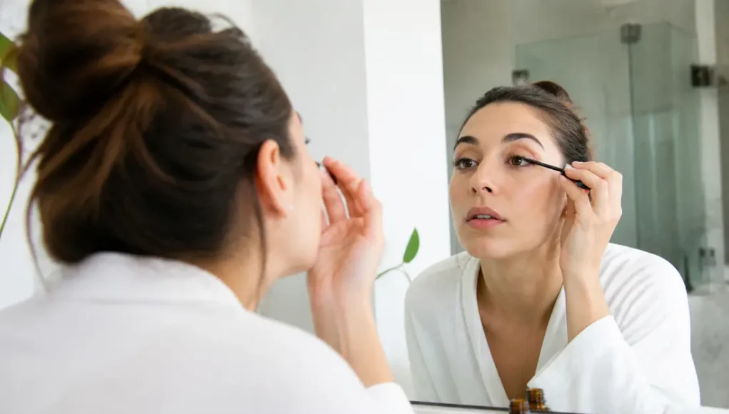 Comment appliquer l'huile de ricin sur les cils sans risque ? Femme appliquant soigneusement de l'huile de ricin cils avec un goupillon propre devant un miroir