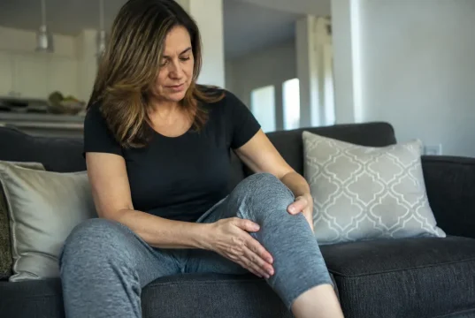 Douleur de la patte d’oie au genou confondue avec le ménisque Femme assise sur un canapé touchant son genou pour soulager une douleur patte d'oie genou.