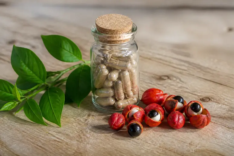 Flacon de complément alimentaire posé à côté de feuilles de thé vert et de graines de guarana sur une table en bois